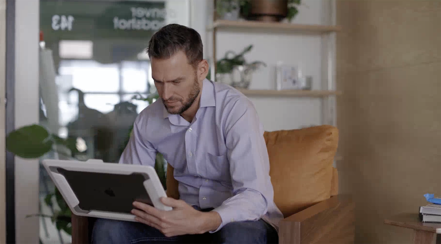 A man sits in an armchair, looking at a tablet device in his hands—perhaps browsing individual headshots—with shelves and plants visible in the background.
